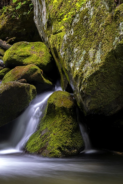 A stream of water made silky smooth with long time exposure photography peacefully flows over mossy rocks.