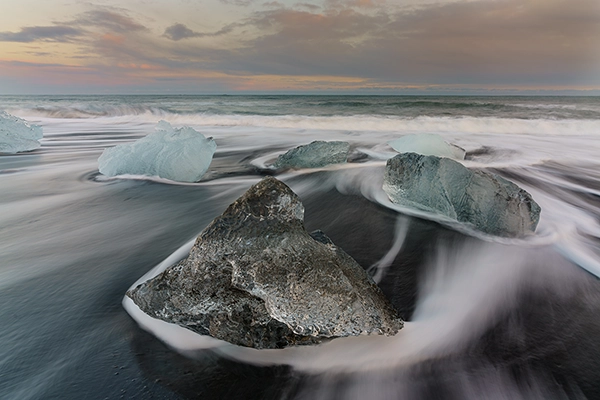 The shore of a black sand beach with large rocks in the foreground photographed using long time exposure photography to make the waves appear smooth.