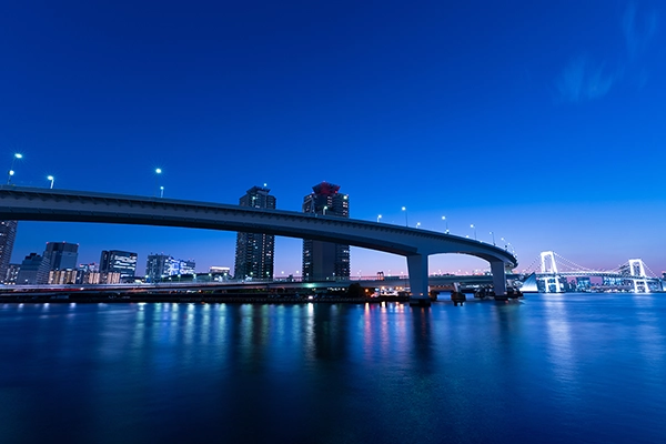 A cityscape featuring a long curved bridge illuminated by street lights photographed at dusk using long exposure photography.