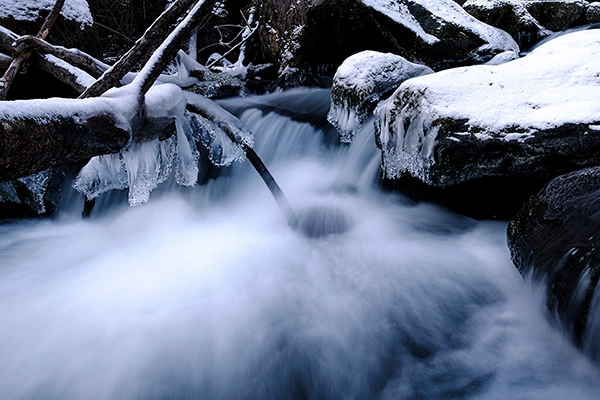 A winter scene showing a stream of water surrounded by trees and rocks partially covered in ice and snow using long time exposure photography techniques. 