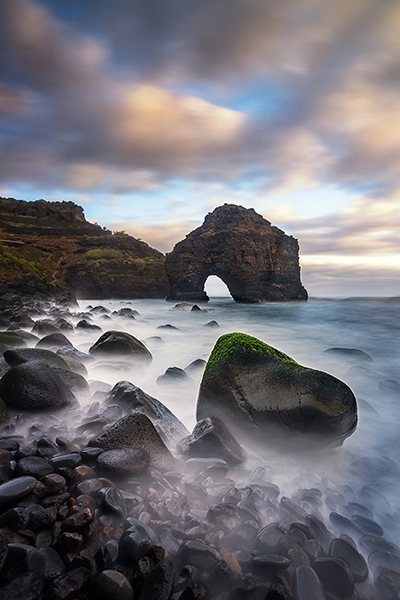 A dramatic shot of a rocky coastline  featuring a natural rock arch formation rising from the sea taken using long time exposure photography to make the water appear more like fog.